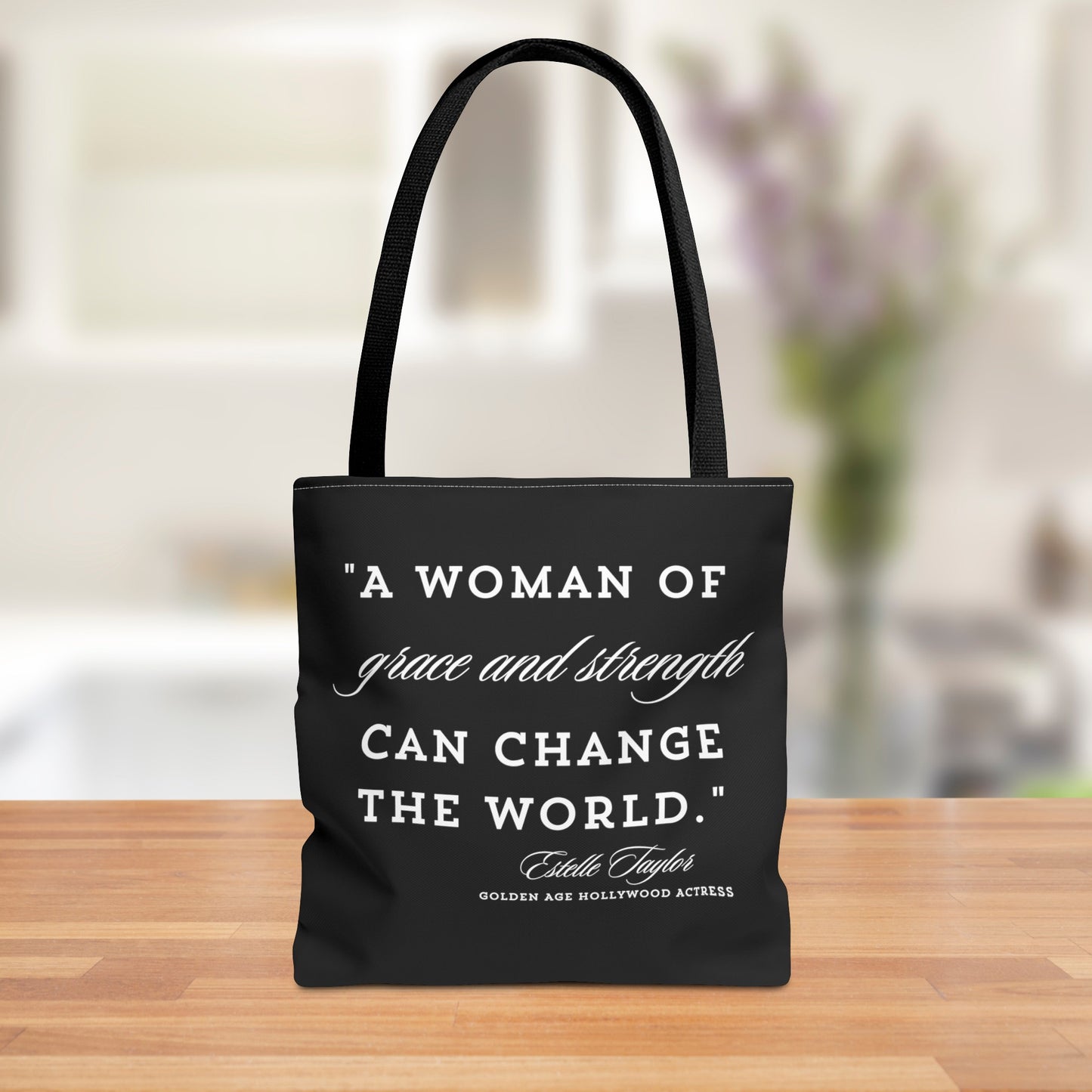 Black tote bag featuring an inspiring quote by Golden Age Hollywood actress Estelle Taylor, stating "A woman of grace and strength can change the world." The white text is displayed in a mix of elegant and bold fonts, offering both a stylish and empowering accessory for women.