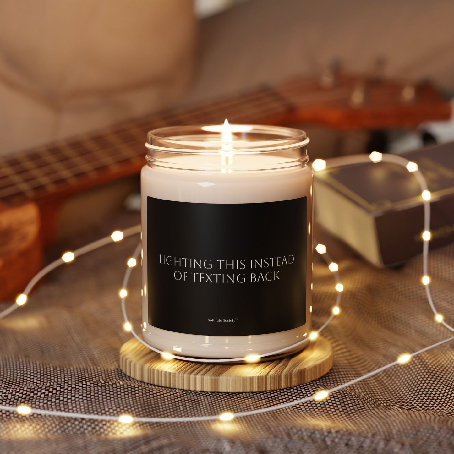 Candle with a humorous message on a wooden coaster surrounded by string lights and a guitar in the background.