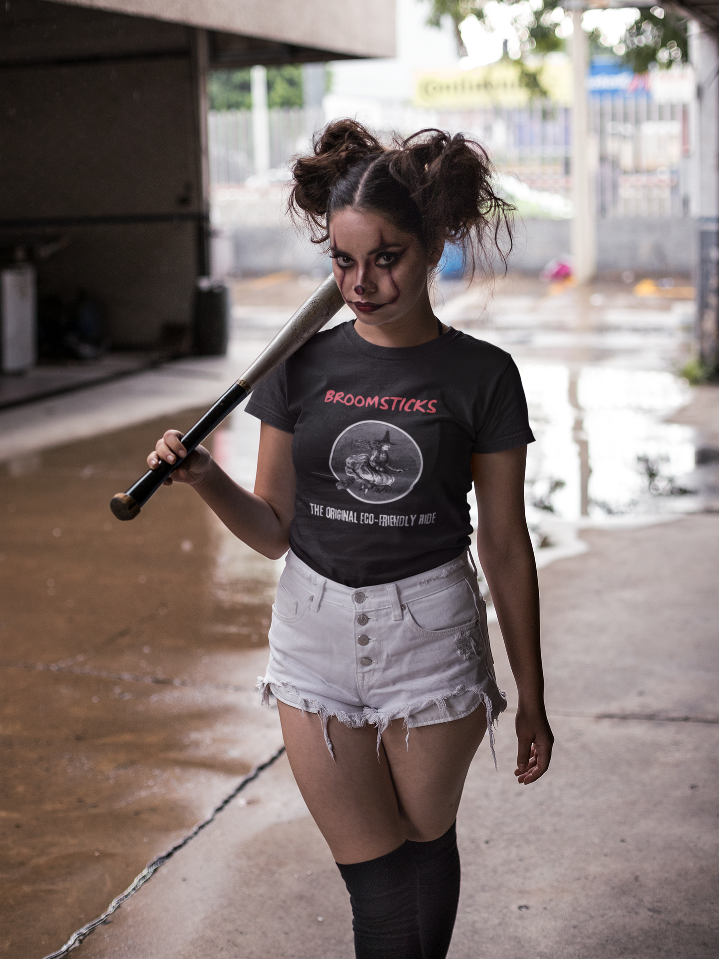 Halloween-themed t-shirt featuring vintage witch design. Young woman dressed in spooky clown makeup and shorts, holding a bat, wearing eco-friendly 'Broomsticks' tee. Perfect for Halloween enthusiasts and eco-conscious shoppers.
