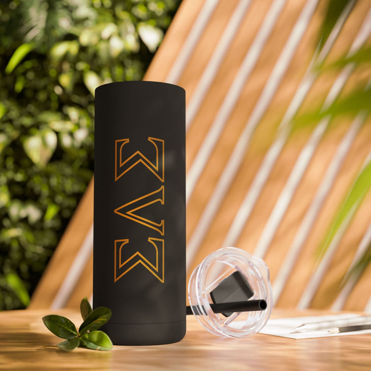 Matte black Soft Life Society skinny tumbler with gold Greek-style design, displayed outdoors on a wooden table with greenery.