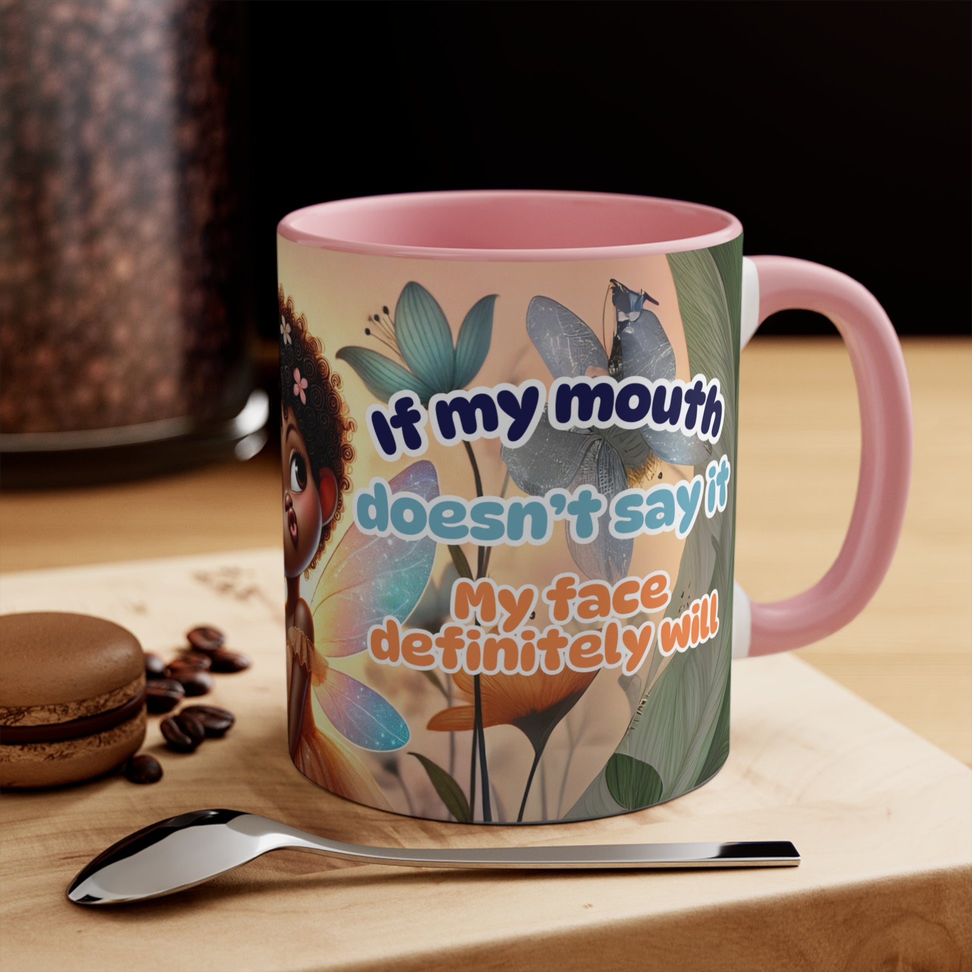 Afro fairy coffee mug with humorous text displayed on a wooden surface with a spoon and macarons

