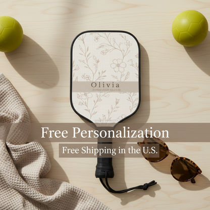 Pickleball paddle with floral design on a wooden surface with balls and sunglasses.