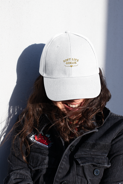 Woman smiling in sunlight while wearing a white “Soft Life Girlie” dad cap and a black denim jacket with embroidered detail.
