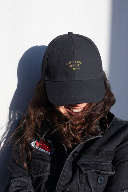 Woman smiling in sunlight while wearing a black “Soft Life Girlie” dad cap and a black denim jacket with embroidered detail.