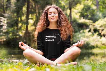 Close-up outdoor meditation scene of a woman sitting cross-legged in nature wearing a black “becoming my best self” sweatshirt.