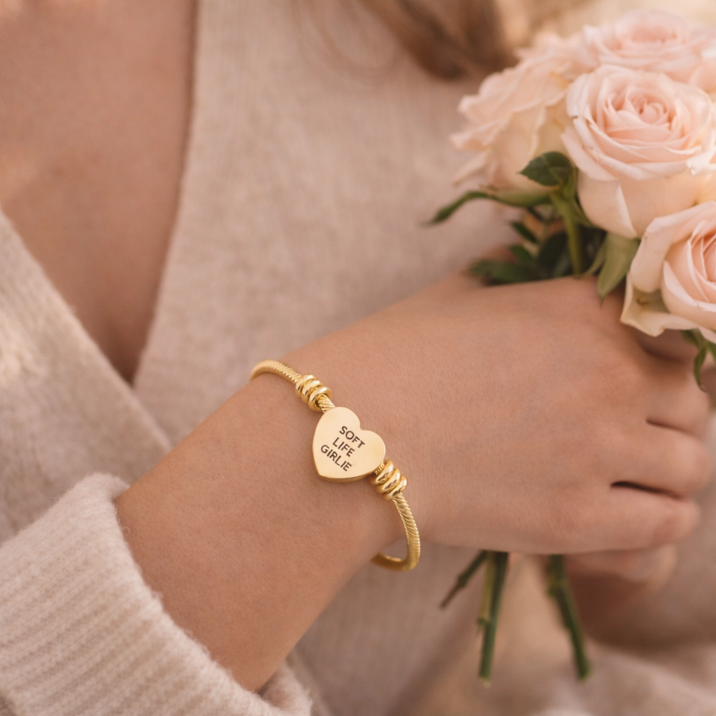 Gold bracelet with heart-shaped charm with engraved text “Soft Life Girlie” worn by a person holding pink roses.