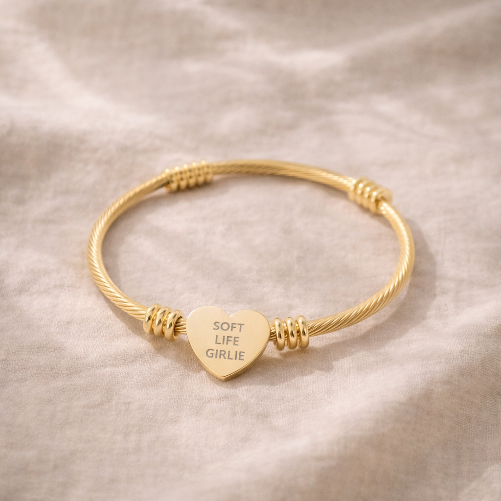 Gold bracelet with a heart-shaped charm with engraved text “Soft Life Girlie” on a beige fabric background