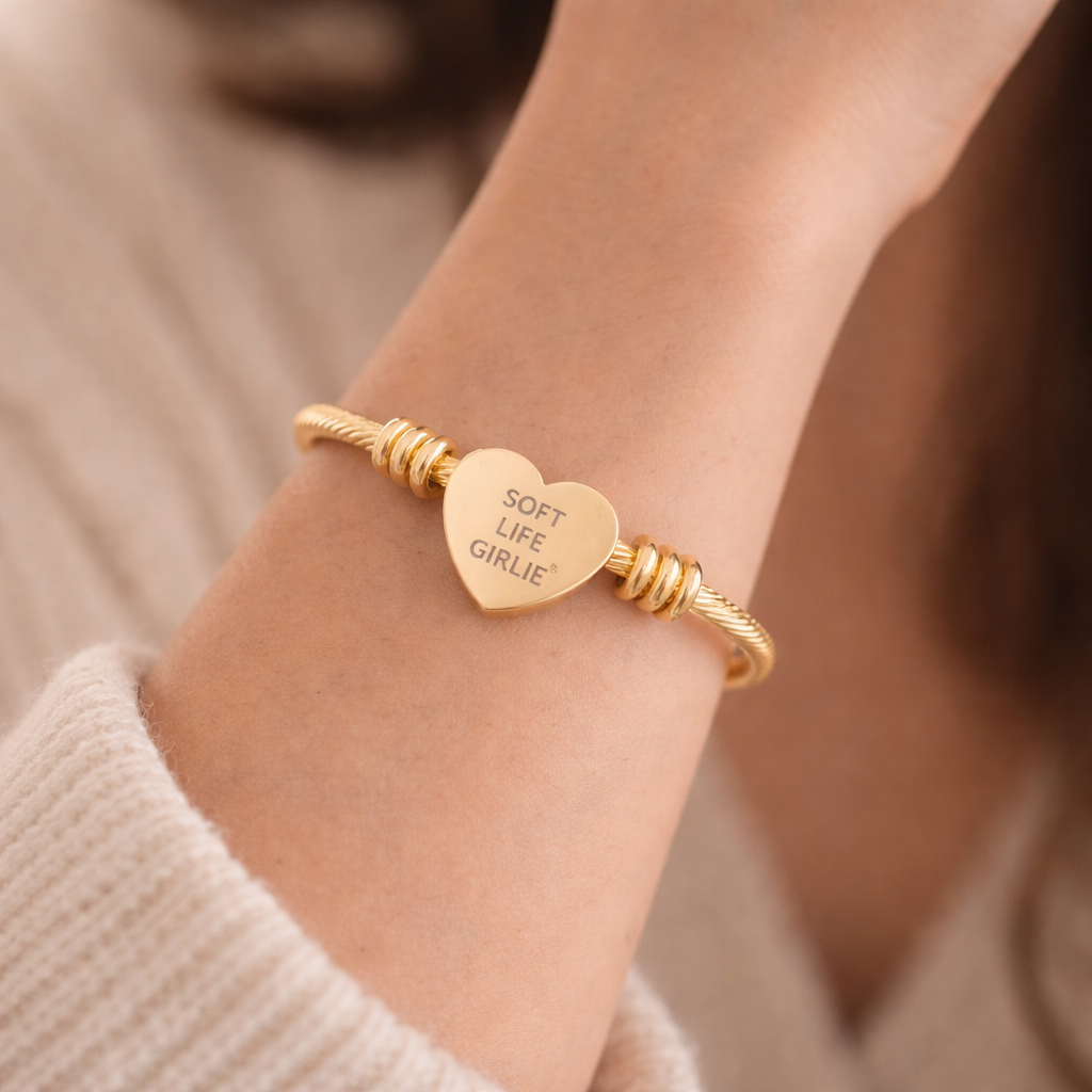 Gold bracelet with a heart-shaped tag with engraved text “Soft Life Girlie” on a wrist, against a neutral background