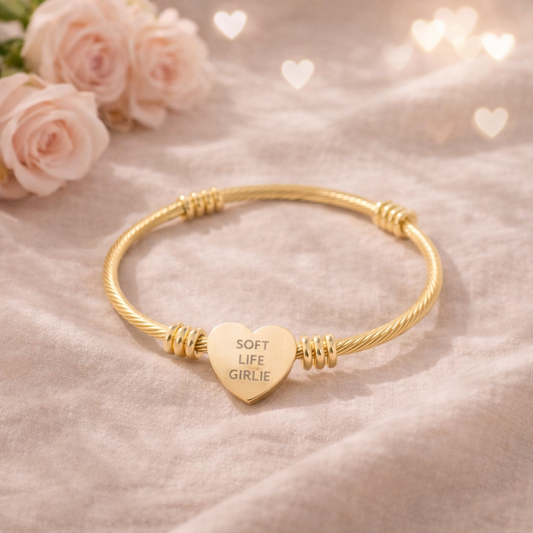 Gold bracelet with a heart-shaped tag with engraved text “Soft Life Girlie” on a soft pink background with roses.