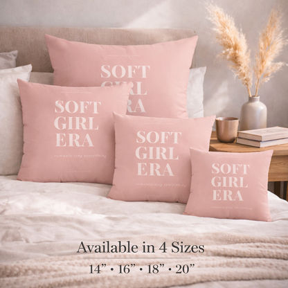 Pink pillows with 'SOFT GIRL ERA' text on a bed, with a side table and decor items in the background.