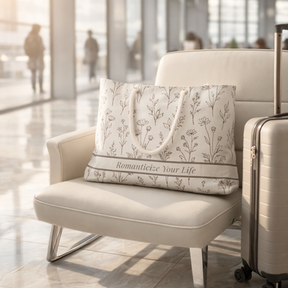 Floral botanical weekender travel bag with rope handles and printed phrase “Romanticize Your Life” resting on airport lounge chair beside suitcase