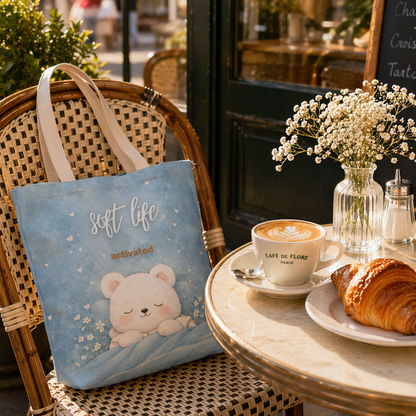 Blue tote bag with a bear design on a chair next to a table with coffee and croissant.
