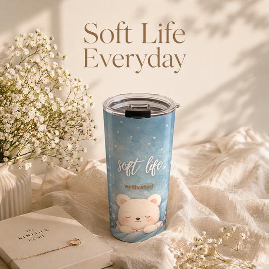 Blue tumbler with a bear design and 'soft life activated' text on a soft fabric background with flowers and a book.