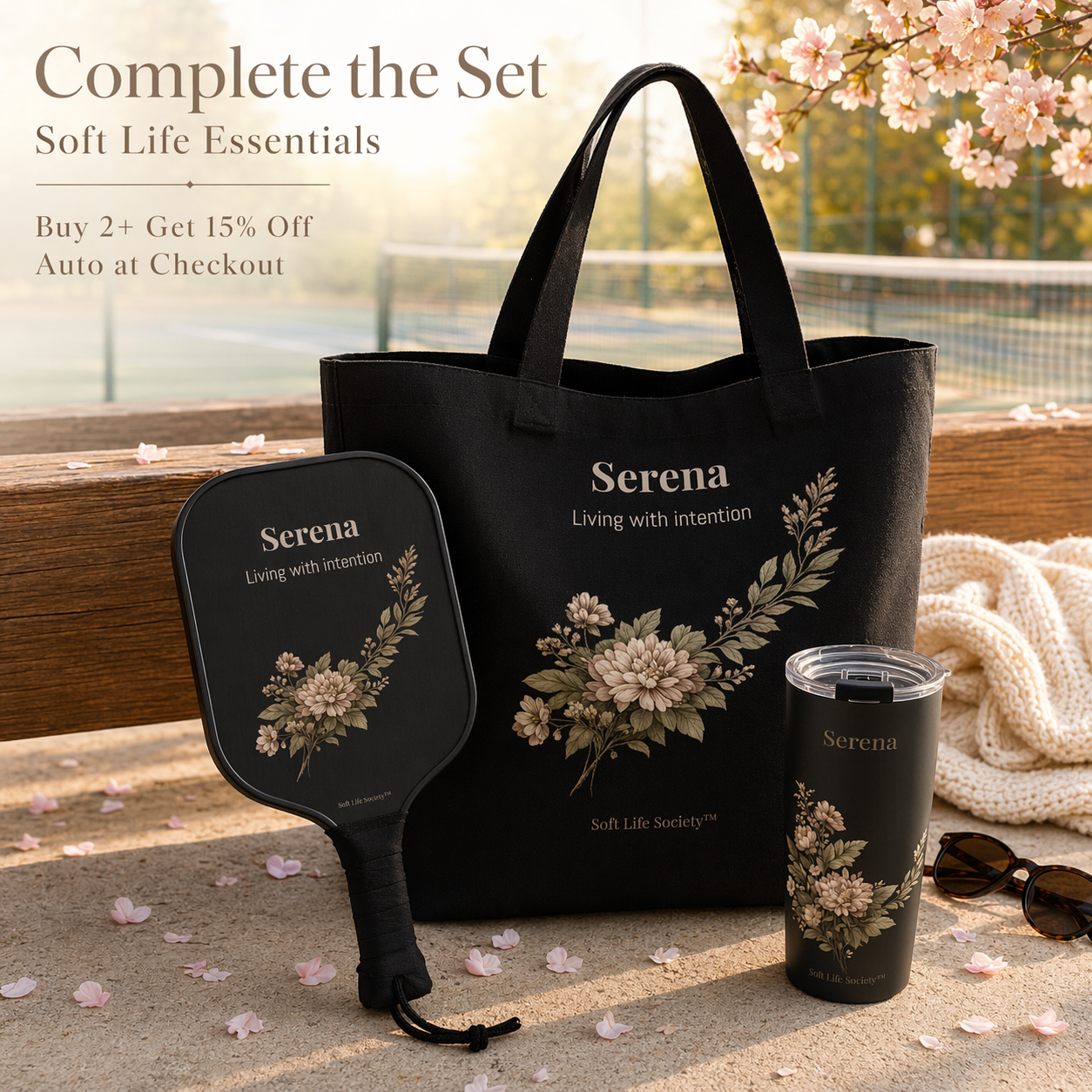 Black tote bag, paddle, and tumbler with floral design and 'Serena' branding on a wooden surface with cherry blossoms.