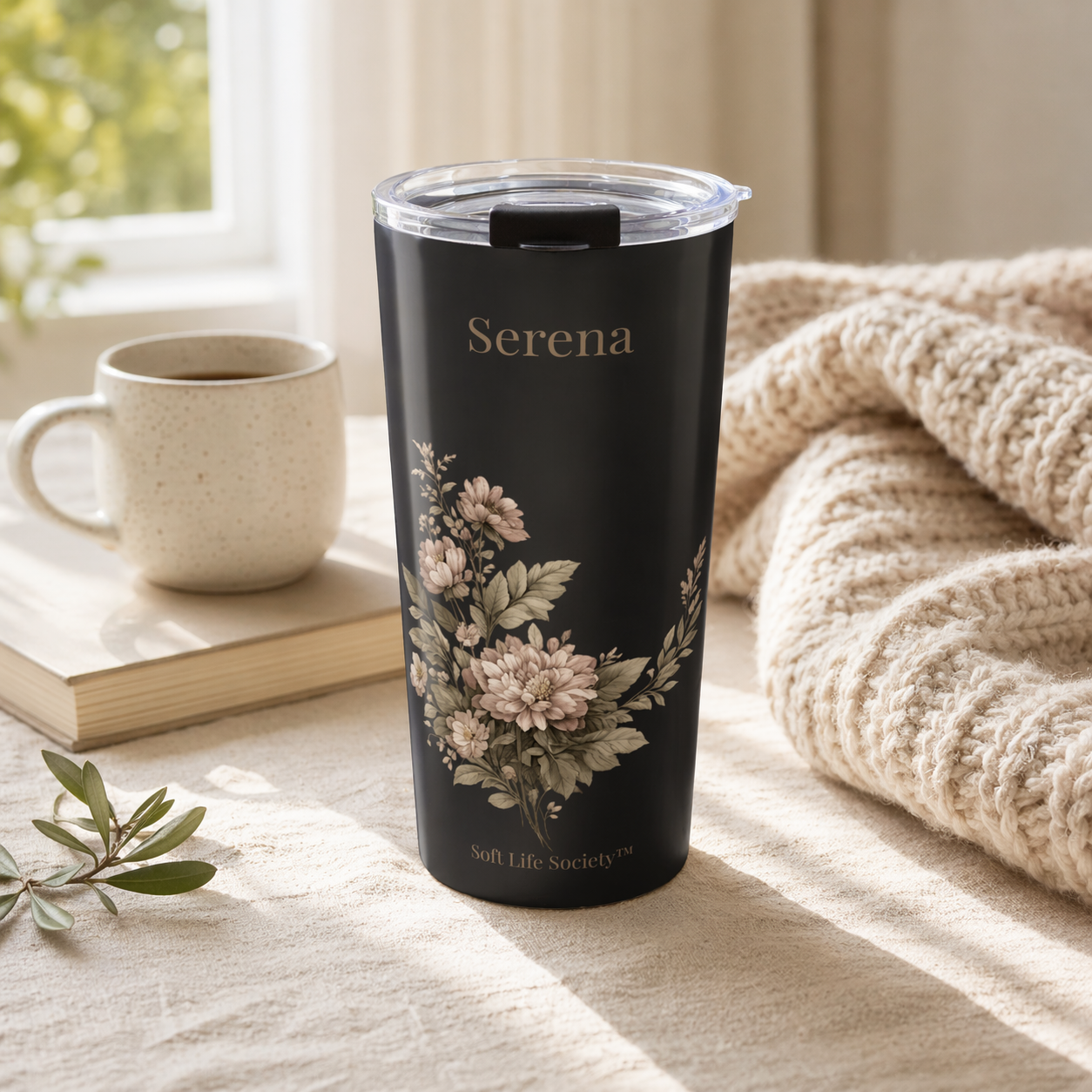 Black floral tumbler with 'Serena' branding on a table with a mug and book in a cozy setting.