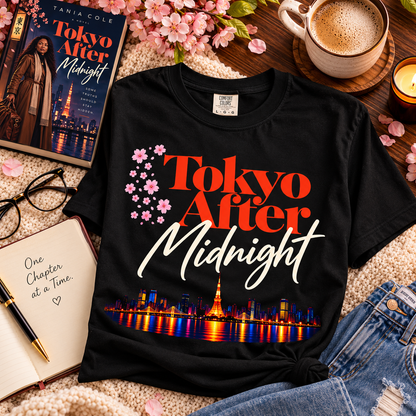 A black graphic T-shirt laid flat on a cozy styled surface with a coffee cup, book, notebook, glasses, flowers, and denim. The shirt reads 'Tokyo After Midnight' in orange and white lettering, with cherry blossoms and a colorful city skyline graphic near the bottom.