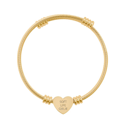 Gold bracelet with a heart-shaped tag with engraved text “Soft Life Girlie” on a white background