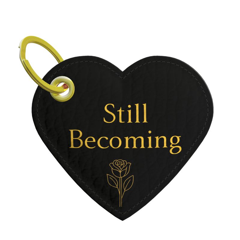 Black heart-shaped keychain with 'Still Becoming' text and a rose design on a white background