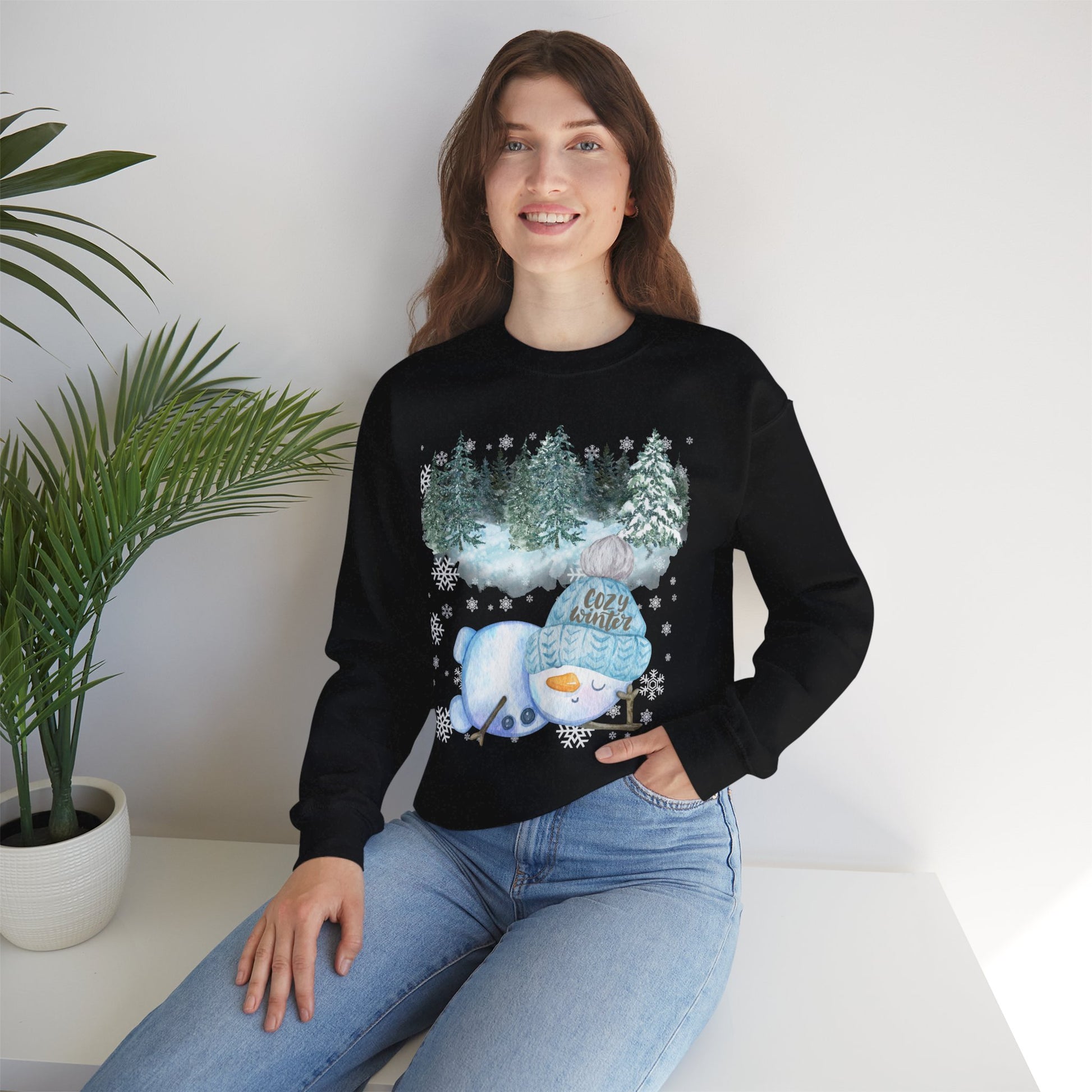 Woman wearing a cozy sweatshirt with a cute sleeping snowman design under a snowy forest, surrounded by snowflakes.