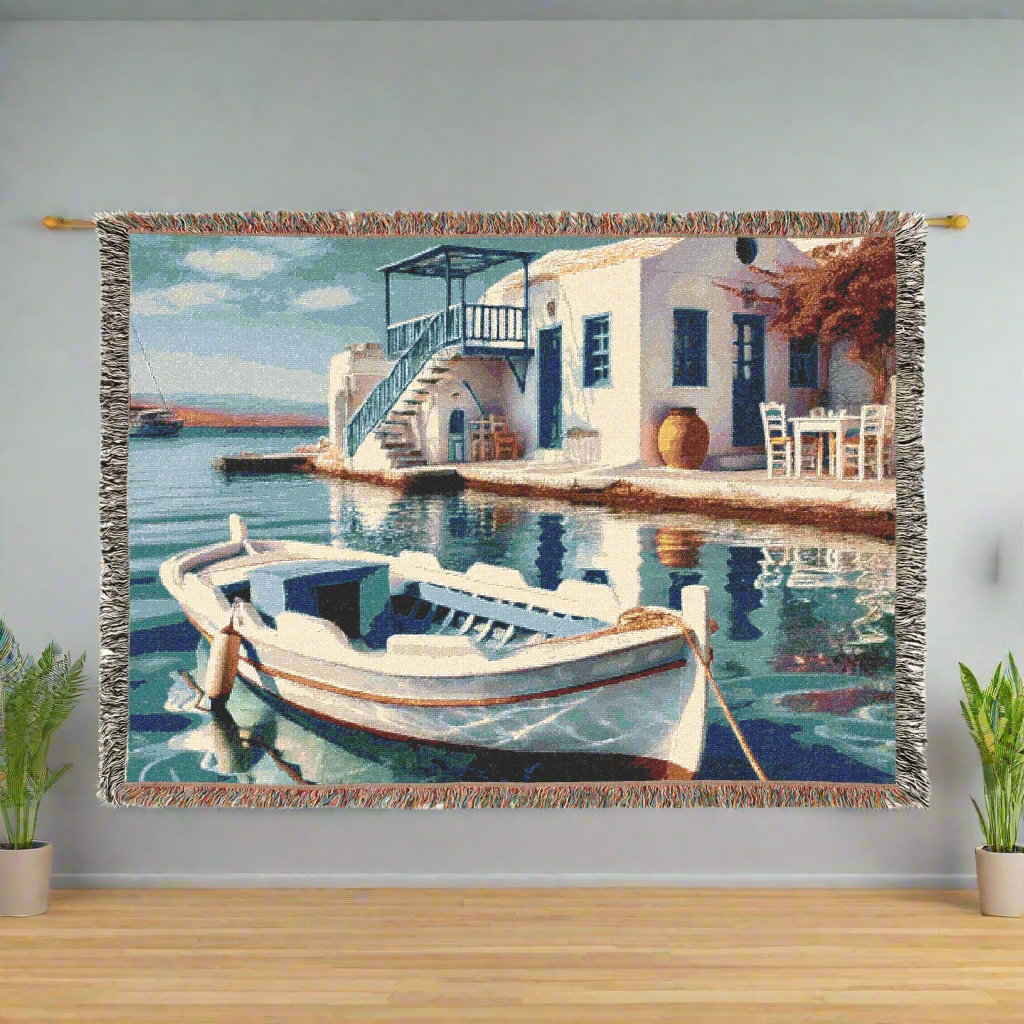 Woven blanket displayed on a wall with a Mediterranean coastal scene featuring a white building, a staircase, and a small boat docked in calm blue waters, surrounded by greenery.