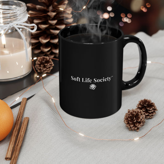 Black 11 oz mug with 'Soft Life Society' logo on a table with decorative items