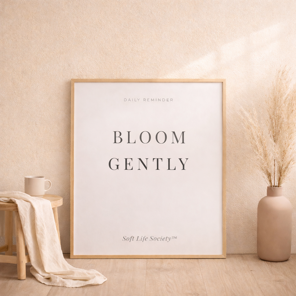 Framed artwork Soft Life Society with 'Bloom Gently' text in a room with a vase and stool.