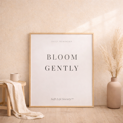 Framed artwork Soft Life Society with 'Bloom Gently' text in a room with a vase and stool.