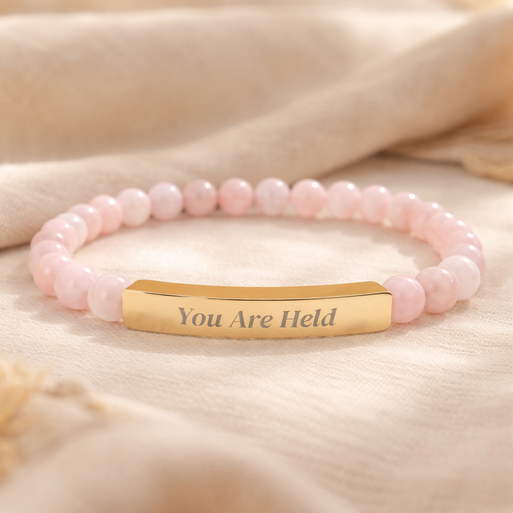 Rose quartz beaded affirmation bracelet with engraved gold bar reading “You Are Held,” photographed on a soft neutral fabric background.
