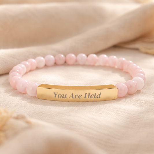 Rose quartz beaded affirmation bracelet with engraved gold bar reading “You Are Held,” photographed on a soft neutral fabric background.