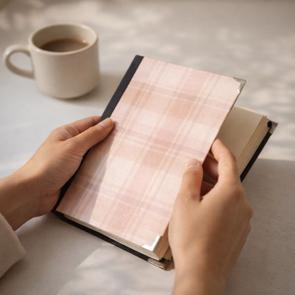 Person holding a notebook with a pink plaid cover on a light surface with a cup of coffee.