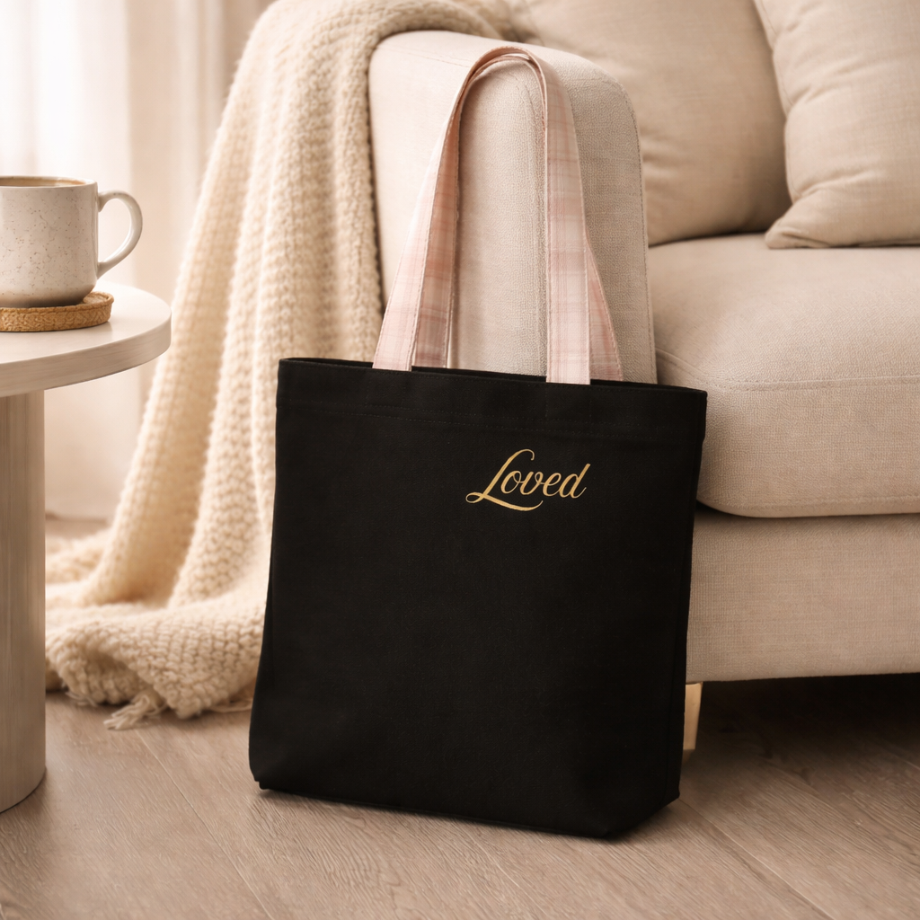 Black tote bag with 'Loved' text in gold on a couch in a cozy living room.