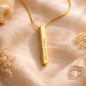 Gold necklace with a vertical pendant on a soft beige fabric background with flowers.