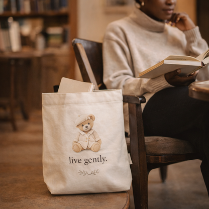 Person reading a book with a tote bag featuring a teddy bear and 'live gently' text in a cozy setting.
