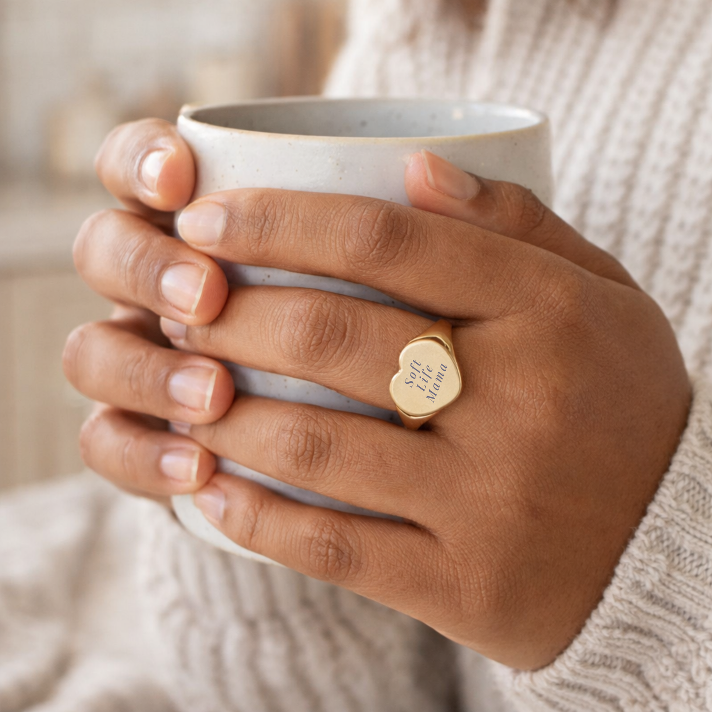 Hand holding a white mug with a gold signet ring with engraving 'Soft Life Mama' on a blurred background