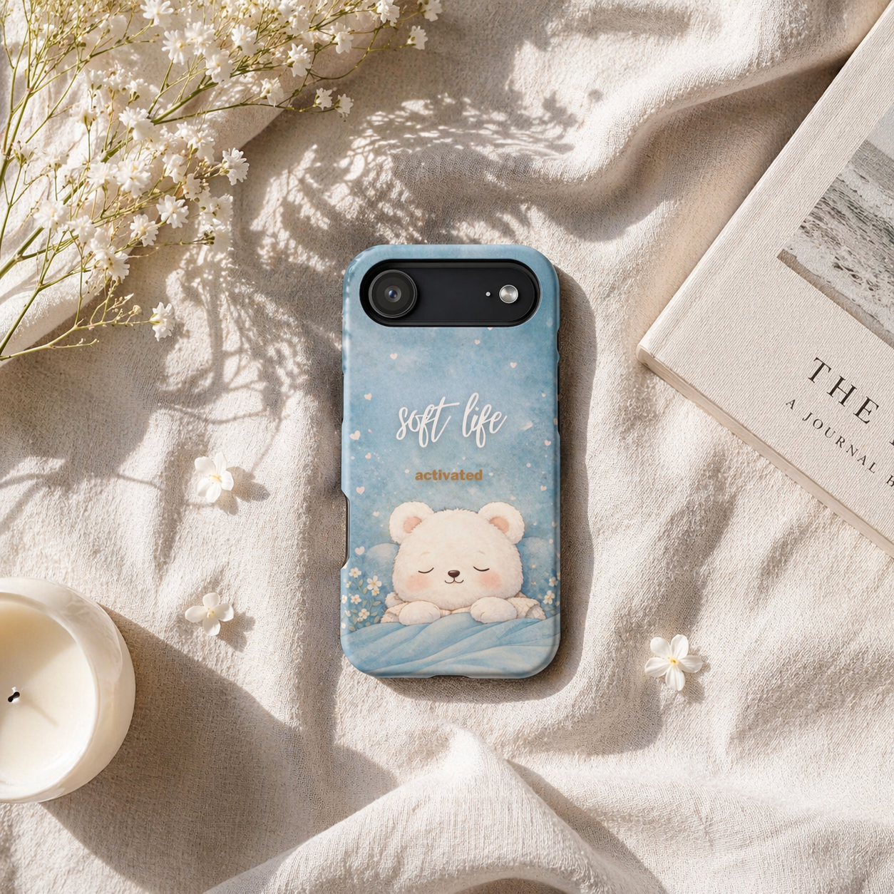 Soft blue bear aesthetic phone case with “Soft Life Activated” text on neutral linen flat lay with flowers and candle