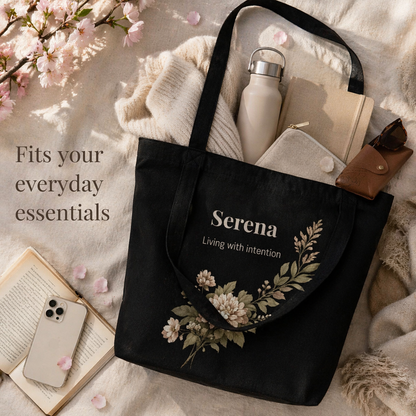 Personalized black canvas tote bag with floral design showing everyday essentials inside neutral aesthetic flat lay