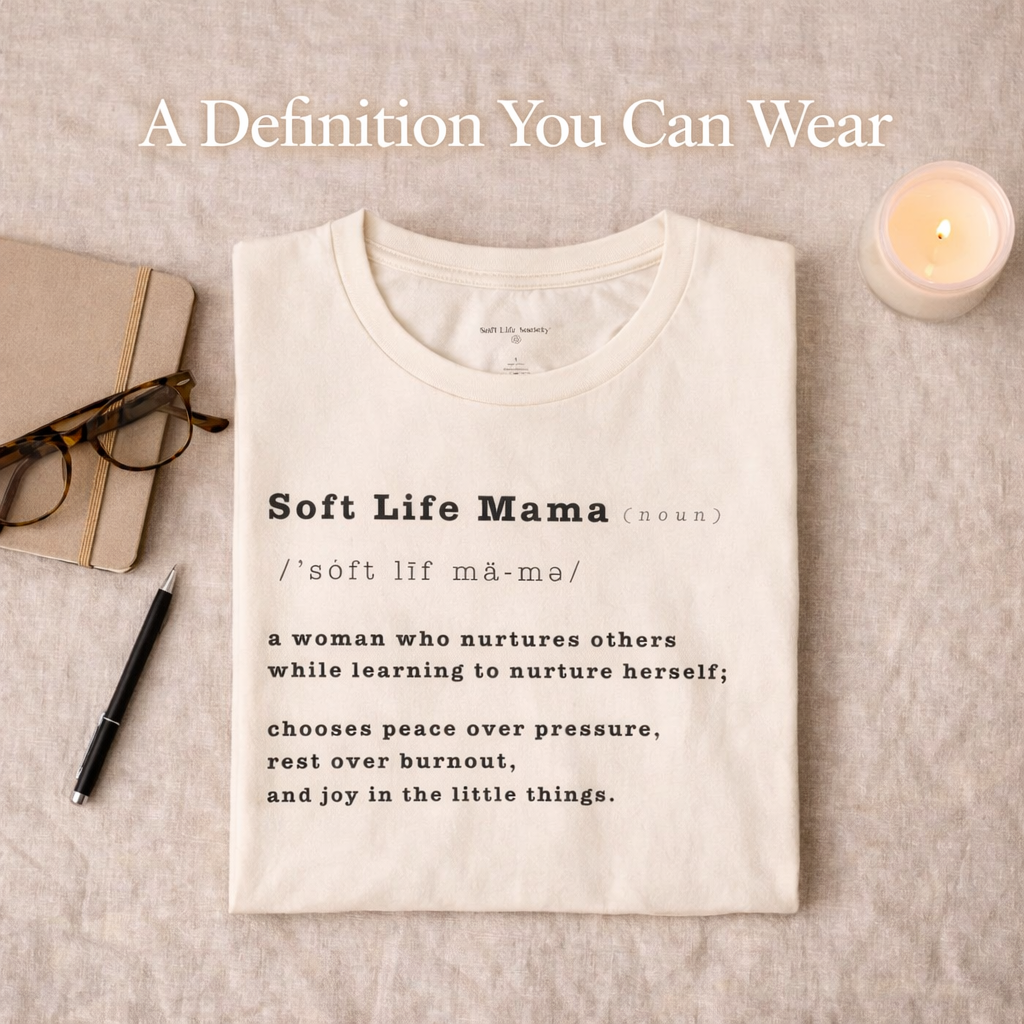 Folded cream-colored t-shirt with 'Soft Life Mama' definition on a textured surface with a notebook, pen, and candle.