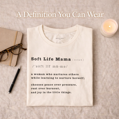 Folded cream-colored t-shirt with 'Soft Life Mama' definition on a textured surface with a notebook, pen, and candle.
