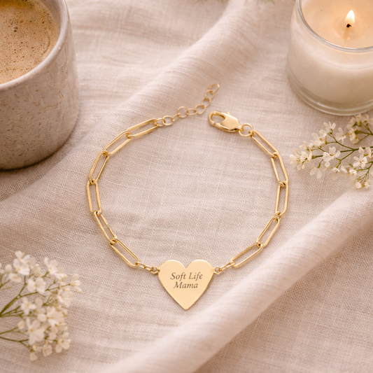 Gold bracelet with heart-shaped pendant engraved 'Soft Life Mama' on a textured surface with a cup of coffee and candle.