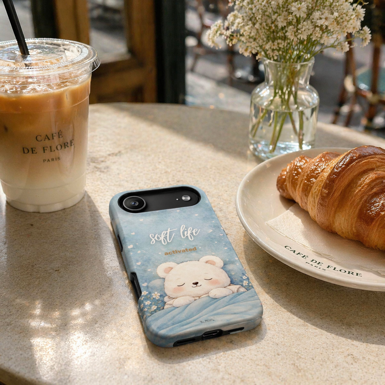 Soft blue “Soft Life Activated” phone case on café table with iced coffee and croissant, everyday aesthetic lifestyle