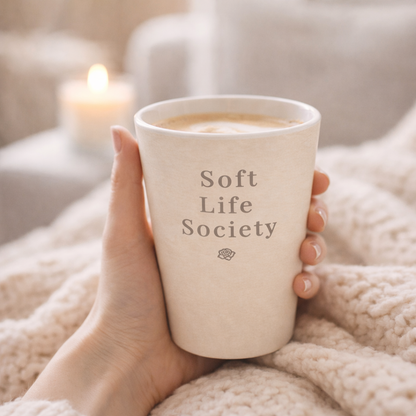 Hand holding a 'Soft Life Society' mug with a blurred candle in the background