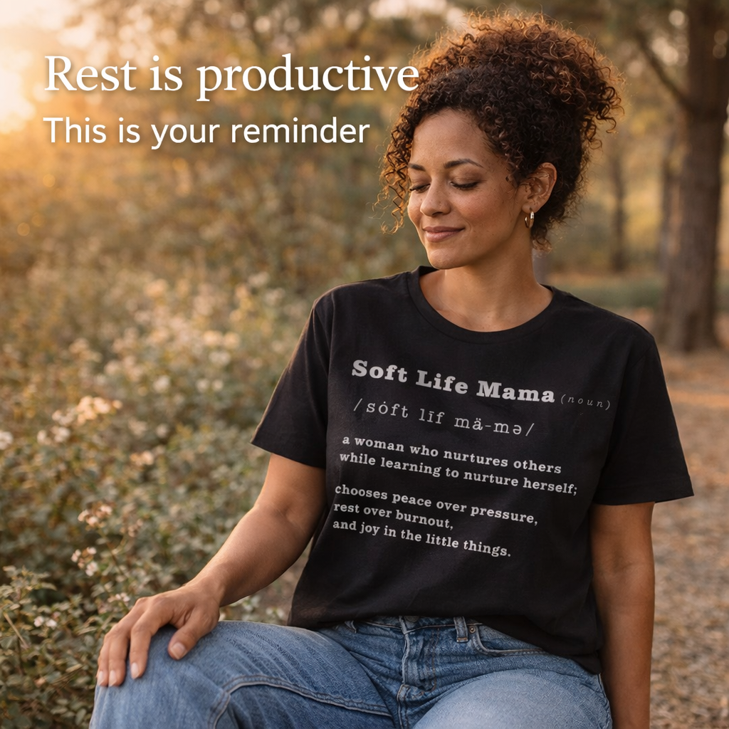 Woman wearing a black t-shirt with 'Soft Life Mama' text, sitting outdoors with nature in the background.