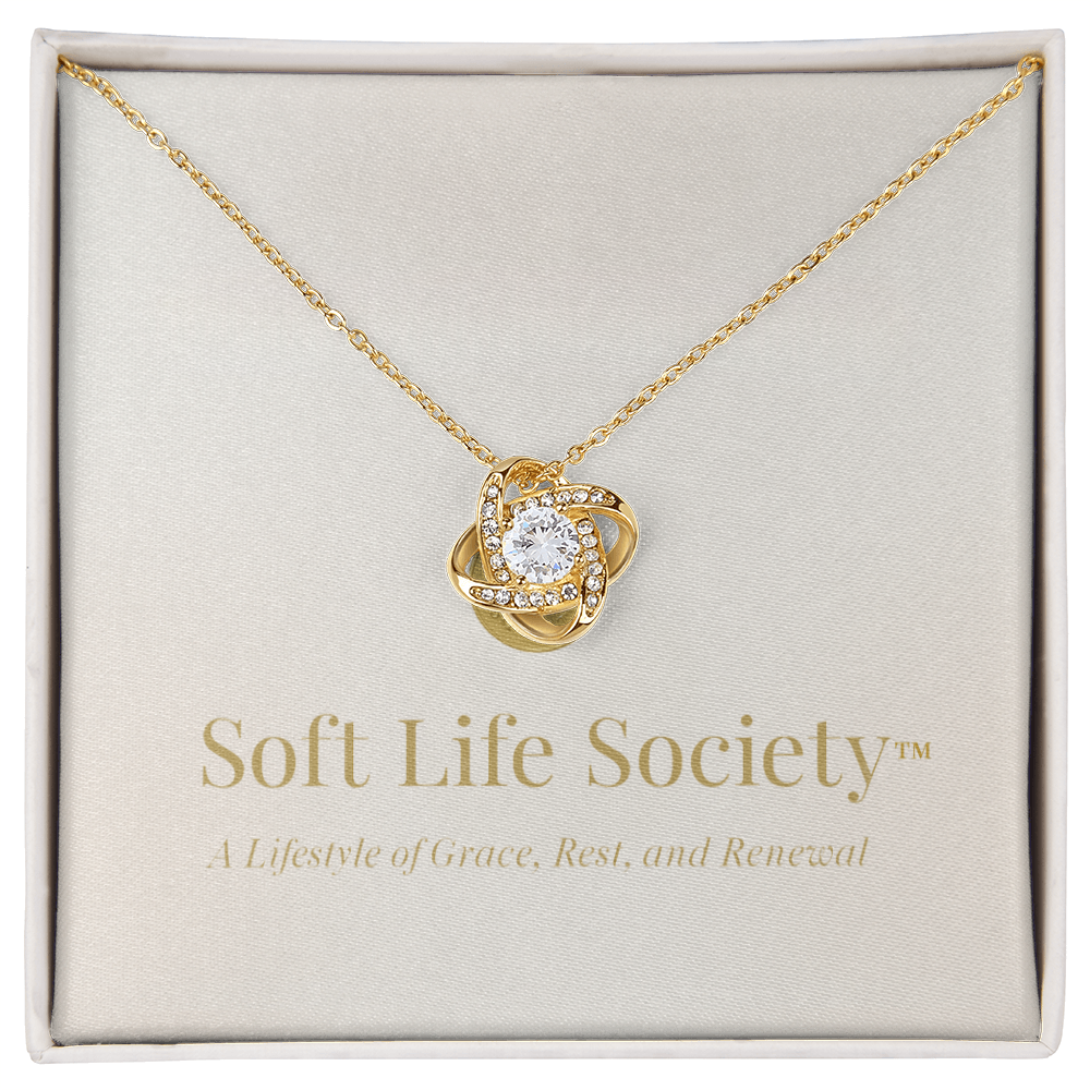 Soft Life Society™ gold Love Knot Necklace displayed in a gift box, featuring a sparkling cubic zirconia pendant on an 18K yellow gold–finished chain above a message card reading ‘A Lifestyle of Grace, Rest, and Renewal.