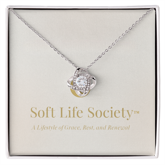 Soft Life Society™ Love Knot Necklace displayed in an elegant cream gift box, featuring a sparkling cubic zirconia center stone and intertwined silver design above the brand’s gold script message card.