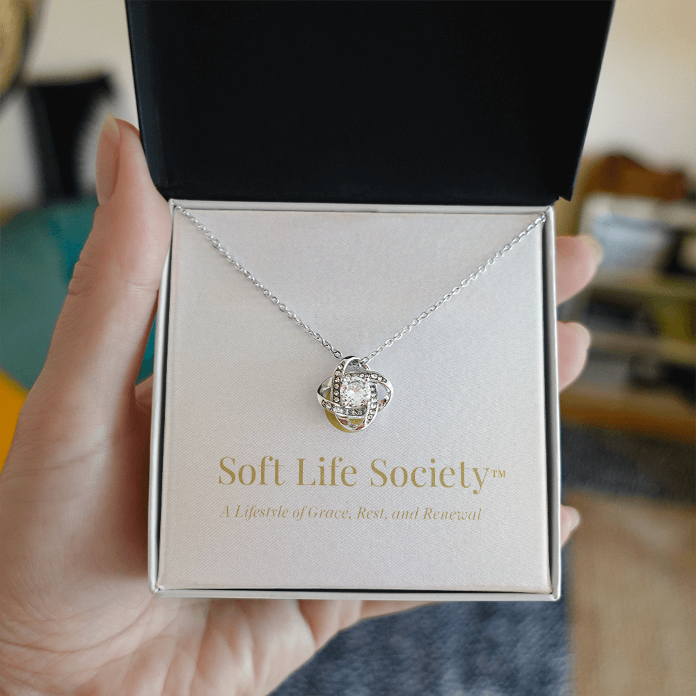 A hand holds an open black jewelry box displaying the Soft Life Society™ Love Knot Necklace, featuring a sparkling cubic zirconia pendant on a silver chain above a message card that reads ‘A Lifestyle of Grace, Rest, and Renewal.’