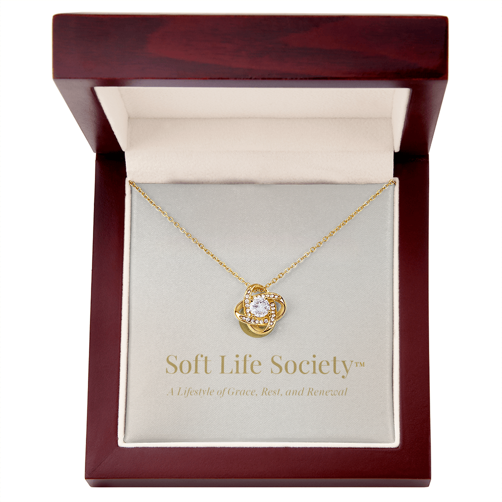 A gold Soft Life Society™ love knot pendant displayed inside a mahogany-style luxury jewelry box, featuring a cubic zirconia center stone and gold-embossed branding that reads “Soft Life Society™ – A Lifestyle of Grace, Rest, and Renewal