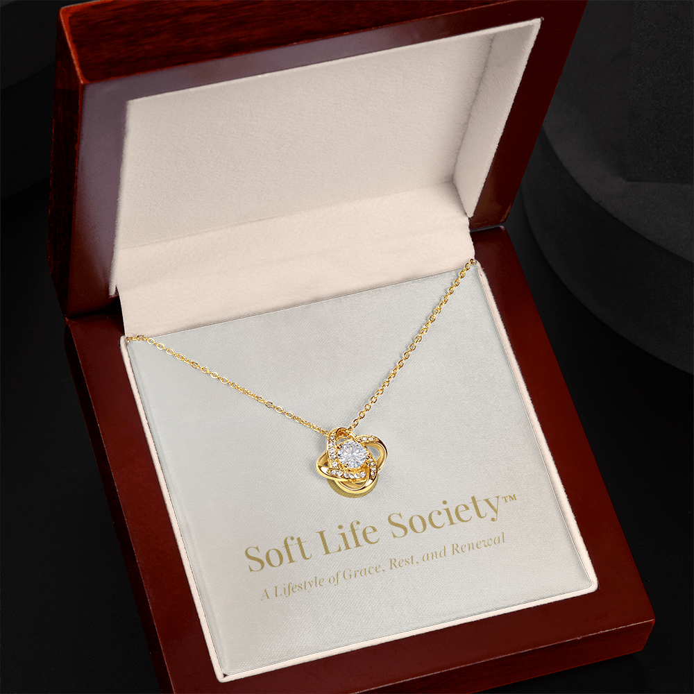 A gold Soft Life Society™ love knot necklace with a sparkling cubic zirconia center displayed inside an open mahogany-style luxury jewelry box, featuring gold-embossed branding that reads “Soft Life Society™ — A Lifestyle of Grace, Rest, and Renewal