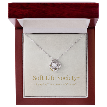 A white gold Soft Life Society™ love knot pendant displayed inside a mahogany-style luxury jewelry box, featuring a cubic zirconia center stone and gold-embossed branding that reads “Soft Life Society™ – A Lifestyle of Grace, Rest, and Renewal.