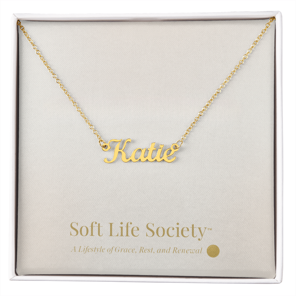 Gold custom name necklace reading ‘Katie’ displayed on a Soft Life Society™ white message card.