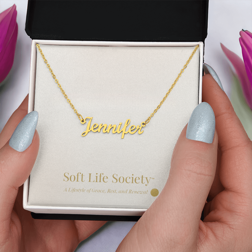 Gold custom name necklace held in hand, presented on a Soft Life Society™ white message card.